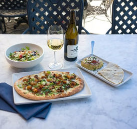 Outdoor patio dining on a marble table: flatbread pizza with roasted vegetables, mixed Mediterranean salad in a bowl, hummus with pita wedges, and a bottle plus glass of white wine.