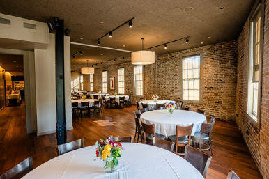 Sunlit industrial-chic event space with exposed brick walls, hardwood floors, large multi-pane windows and round tables covered in white linens with colorful floral centerpieces.