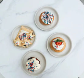 Top-down view of four round plates on white marble holding assorted frosted desserts—cupcakes and star-shaped cookies—topped with whipped frosting, a sliced strawberry, and red, white, and blue sprinkles.