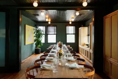 Long communal wooden dining table set for a dinner party in a cozy urban private dining room with dark green walls, round wooden chairs, globe ceiling lights, floral centerpieces, folded napkins and windows.
