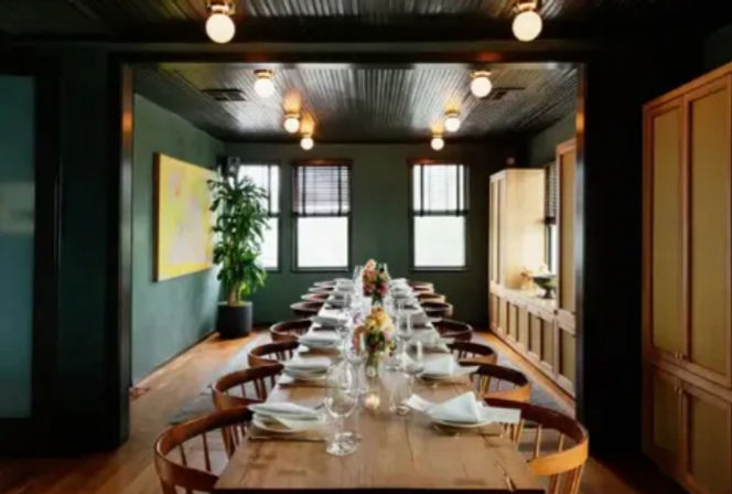 Long communal wooden dining table set for a dinner party in a cozy urban private dining room with dark green walls, round wooden chairs, globe ceiling lights, floral centerpieces, folded napkins and windows.
