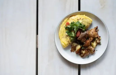 Brunch plate with a folded fluffy omelet topped with cherry tomato and fresh herbs, served with crispy roasted potatoes on a white plate over a light wooden farmhouse table