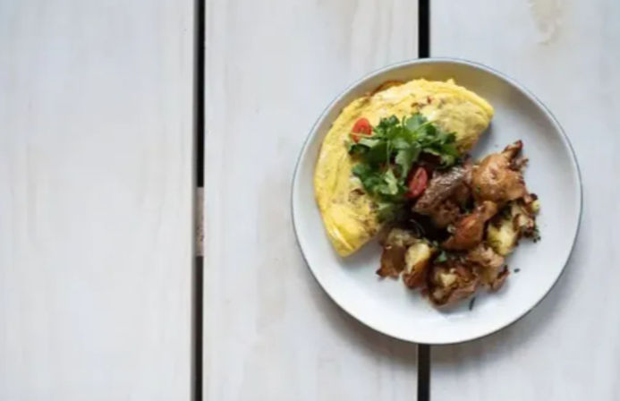 Brunch plate with a folded fluffy omelet topped with cherry tomato and fresh herbs, served with crispy roasted potatoes on a white plate over a light wooden farmhouse table