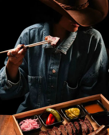 Close-up of a person in a cowboy hat and denim jacket using chopsticks to lift a slice of grilled steak from a wooden tray with sliced beef, pickled onions, peppers and dipping sauces.