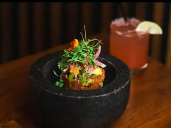 Bright seafood tartare piled in a black stone mortar, topped with microgreens and edible flowers, with a pink lime-garnished cocktail on a wooden table.