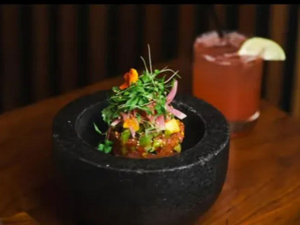 Bright seafood tartare piled in a black stone mortar, topped with microgreens and edible flowers, with a pink lime-garnished cocktail on a wooden table.