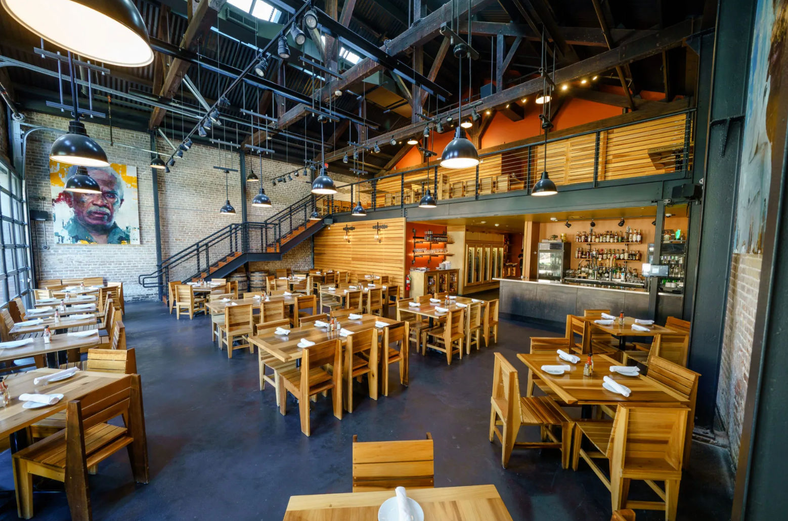 Industrial loft-style restaurant interior with exposed brick walls, open wooden beams and mezzanine, rows of wooden tables set with napkins under large black pendant lights