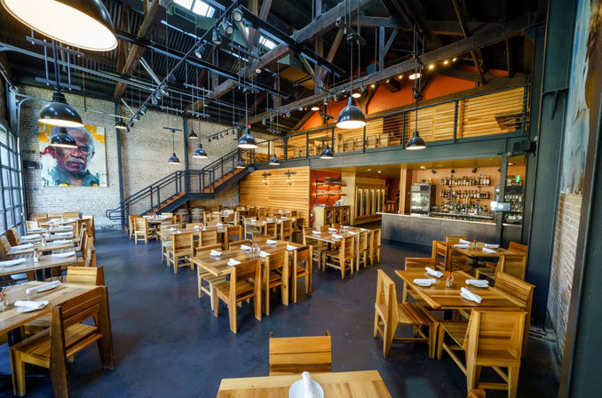 Industrial loft-style restaurant interior with exposed brick walls, open wooden beams and mezzanine, rows of wooden tables set with napkins under large black pendant lights