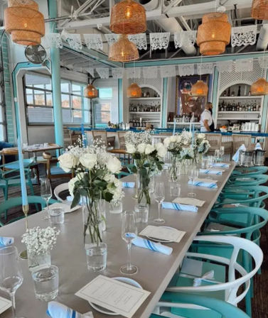 Sunlit coastal-style restaurant interior with a long communal table set for dining, turquoise chairs, vases of white roses, rattan pendant lights, and a staff member at the bar.