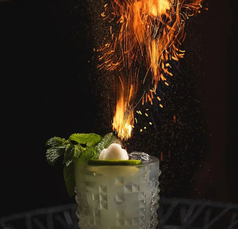 Close-up of a craft cocktail in a textured glass with mint sprig and lime wheel, flaming citrus oil sending dramatic orange sparks and embers into a dark background.