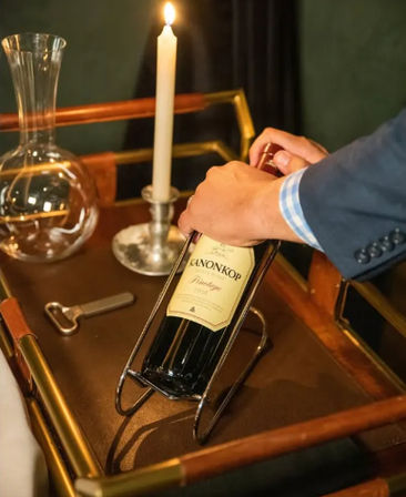 Hand placing a red wine bottle into a metal cradle on a brass-trimmed serving cart beside a lit taper candle and glass decanter — cozy dinner setup.