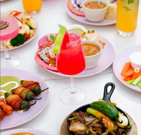 Vibrant Mexican-inspired brunch spread on a white table: bright pink watermelon cocktail garnished with a lime/watermelon wedge, tostada with salsa and rice, bacon-wrapped jalapeños, sizzling skillet fajitas with grilled peppers and onions, and plates of veggies and dips.