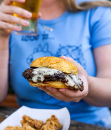 Close-up of a juicy cheeseburger with melted cheese on a golden brioche bun, held by a person sipping a beer; paper tray of fried chicken in the foreground.