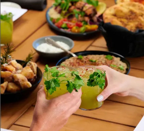 Two hands clinking cilantro-garnished green cocktails over a wooden table surrounded by bowls of fried bites, dips, salad and bread for casual patio dining
