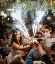 Woman in white holding lit sparklers above a dinner table in a lively indoor restaurant celebration with hanging greenery and twinkling lights