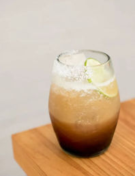 Icy salt-rimmed cocktail in a stemless glass with lime wedge, layered amber-to-brown hues, served on a wooden table.
