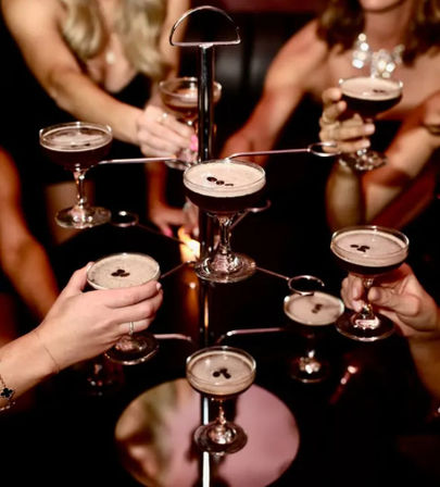Tiered cocktail tower holding espresso martinis in coupe glasses, each with coffee-bean garnish, hands clinking glasses in a dim lounge setting.