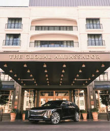 Luxury hotel entrance with a wide canopy and hanging marquee lights, balconies and glass doors above, potted plants at the curb and a sleek black SUV parked out front.