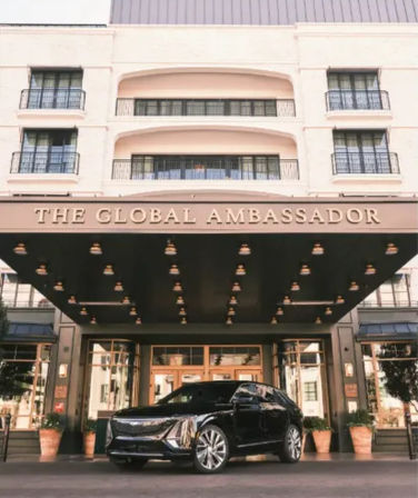 Luxury hotel entrance with a wide canopy and hanging marquee lights, balconies and glass doors above, potted plants at the curb and a sleek black SUV parked out front.