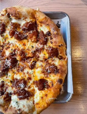 Close-up of a cheesy pizza topped with browned sausage crumbles and grated Parmesan, served on a metal tray on a light wooden restaurant table.