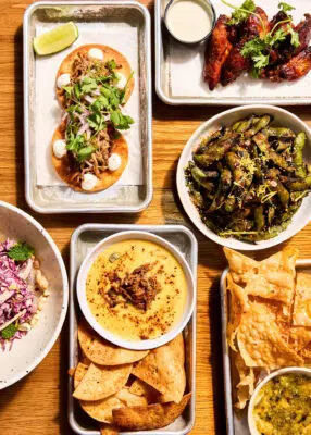 Overhead shot of a vibrant casual-food spread on a wooden table: two cilantro-topped tacos with lime, spicy chicken wings, seasoned edamame, tortilla chips with creamy dip, and a fresh slaw.