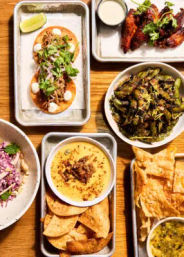 Overhead shot of a vibrant casual-food spread on a wooden table: two cilantro-topped tacos with lime, spicy chicken wings, seasoned edamame, tortilla chips with creamy dip, and a fresh slaw.