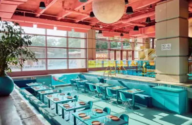 Turquoise indoor pool converted into a retro dining area with tables and chairs set in the shallow end, large floor-to-ceiling windows, pink ceiling and a visible "No Diving" sign.