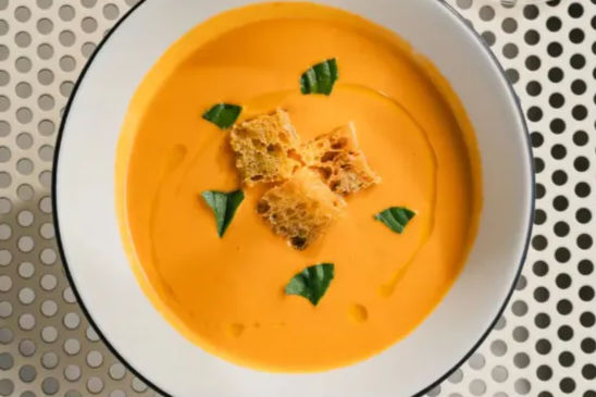 Overhead view of a bright creamy orange soup garnished with crunchy croutons and basil leaves in a white bowl on a perforated metal surface.