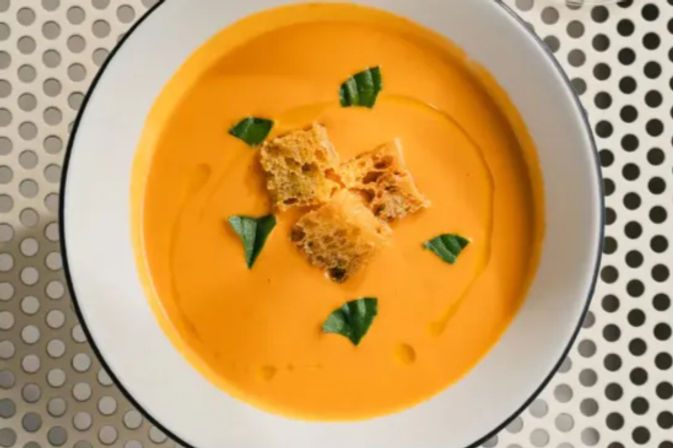 Overhead view of a bright creamy orange soup garnished with crunchy croutons and basil leaves in a white bowl on a perforated metal surface.