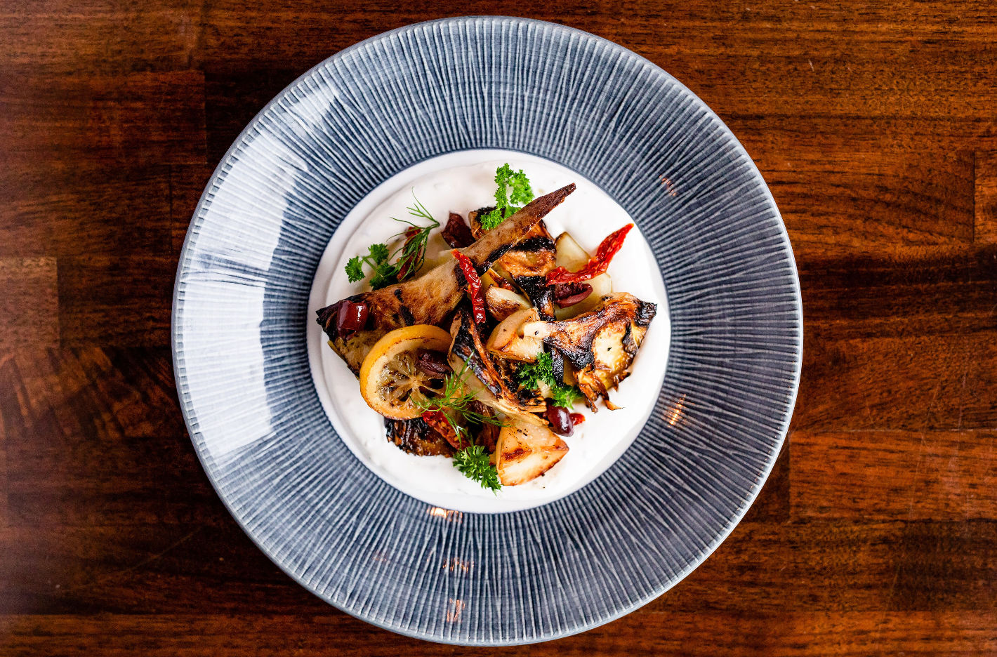 Top-down view of a gourmet roasted vegetable medley with grilled lemon, dried red chilies and fresh herbs on a creamy white sauce, plated on a textured blue-rimmed plate over a wooden table