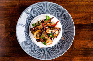Top-down view of a gourmet roasted vegetable medley with grilled lemon, dried red chilies and fresh herbs on a creamy white sauce, plated on a textured blue-rimmed plate over a wooden table