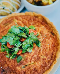 Golden-brown savory quiche on a white plate topped with fresh cilantro, halved cherry tomatoes and mixed greens — brunch-style close-up