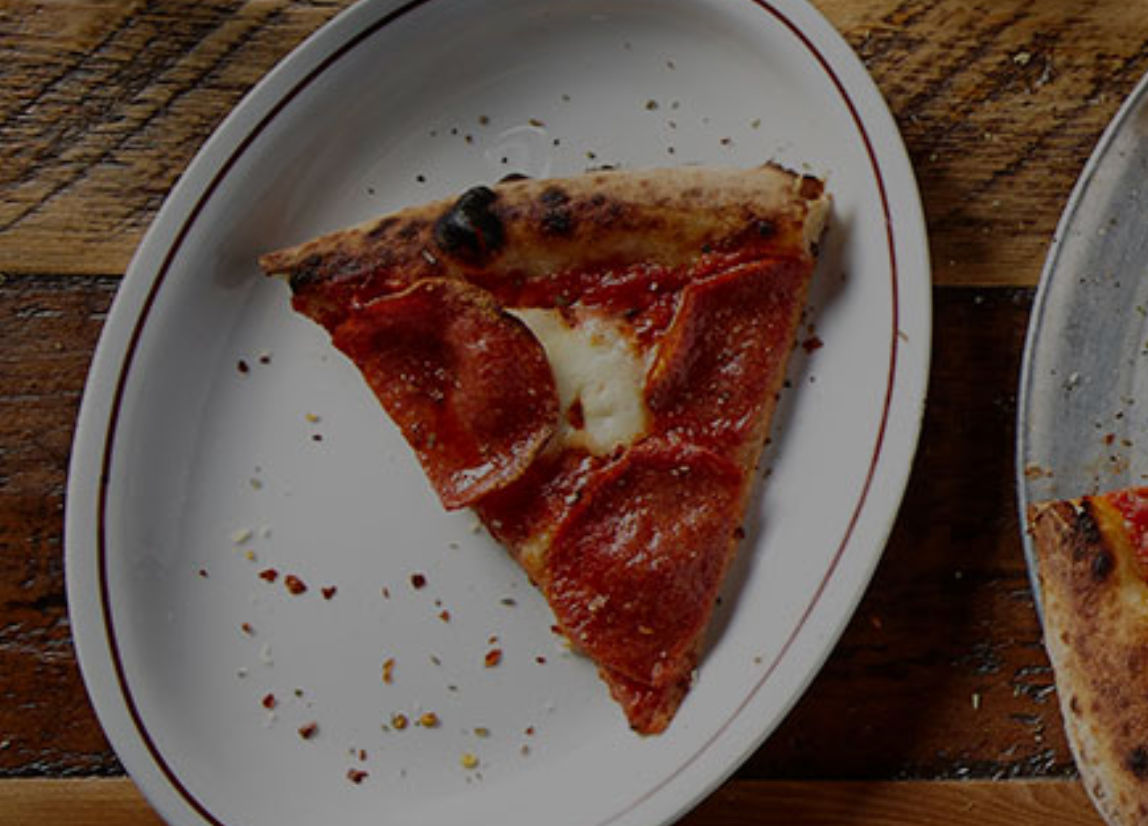 Single pepperoni pizza slice with melted mozzarella on a white oval plate with a red rim, sprinkled with red pepper flakes on a rustic wooden table