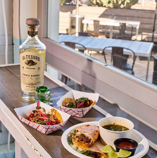 Sunlit Mexican spread on a windowsill: two loaded street tacos in paper trays, a grilled quesadilla with meat, lime wedges, bowls of salsa and green soup, and a tequila bottle with a shot glass, outdoor patio visible.