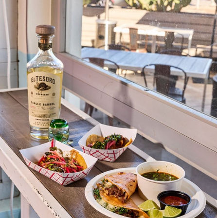 Sunlit Mexican spread on a windowsill: two loaded street tacos in paper trays, a grilled quesadilla with meat, lime wedges, bowls of salsa and green soup, and a tequila bottle with a shot glass, outdoor patio visible.
