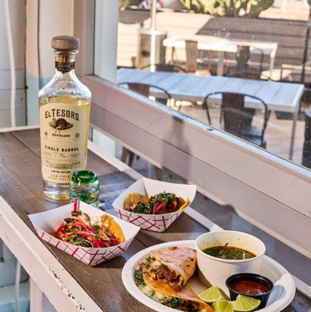 Sunlit Mexican spread on a windowsill: two loaded street tacos in paper trays, a grilled quesadilla with meat, lime wedges, bowls of salsa and green soup, and a tequila bottle with a shot glass, outdoor patio visible.