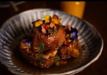 Glazed braised pork belly medallions topped with bright fruit salsa, mint and edible flowers served in a shallow blue ceramic bowl