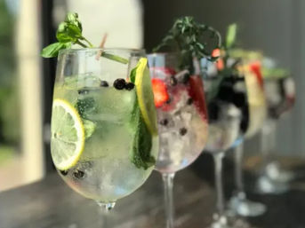 Row of refreshing summer cocktails in stemmed wine glasses on a patio bar, filled with ice, lemon and cucumber slices, berries and fresh herbs