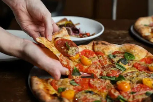 Hand pulling a slice of wood-fired pizza topped with fresh basil, cherry tomatoes, tomato slices and savory meat on a rustic table