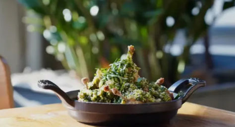 Cast-iron skillet piled with sautéed greens, herbs and crispy crumbs on a wooden table with blurred outdoor patio greenery