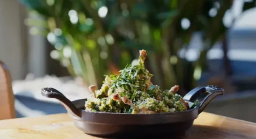 Cast-iron skillet piled with sautéed greens, herbs and crispy crumbs on a wooden table with blurred outdoor patio greenery