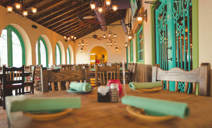 Rustic Mexican-style dining room with arched windows, teal wrought-iron doors, wooden tables set with green napkins and warm chandeliers under exposed beam ceiling.