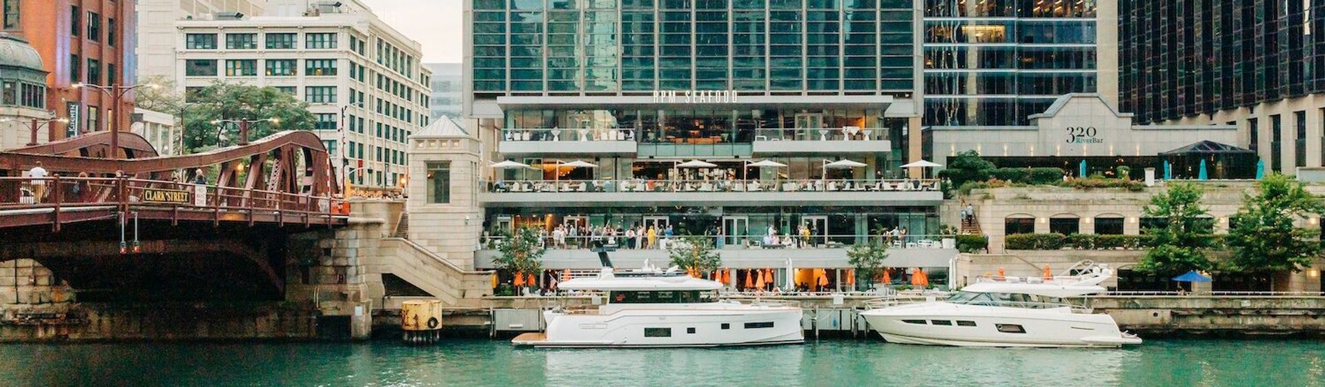 Two white yachts moored on the Chicago River beside a bustling multi-level riverwalk with outdoor dining, glass high-rises and a red steel bridge.