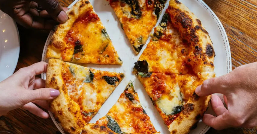 Overhead view of a wood-fired margherita pizza on a white plate with bubbling mozzarella, tomato sauce and basil, three hands reaching to grab slices from a rustic wooden table.