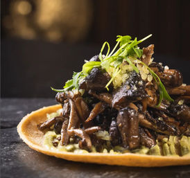 Close-up of a gourmet sautéed mushroom taco on a toasted corn tortilla with avocado crema and microgreens on a dark stone background.