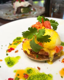 Close-up of a vibrant brunch eggs Benedict on a toasted English muffin with a poached egg, glossy hollandaise, diced tomatoes and fresh cilantro, plated with green herb oil drizzles and chive garnish.