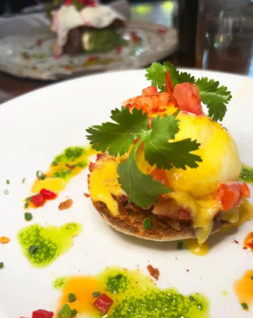Close-up of a vibrant brunch eggs Benedict on a toasted English muffin with a poached egg, glossy hollandaise, diced tomatoes and fresh cilantro, plated with green herb oil drizzles and chive garnish.