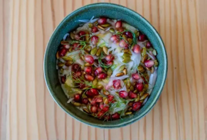 Top-down view of a green bowl with creamy yogurt topped with pomegranate seeds, pepitas and fresh fennel fronds on a light wooden table