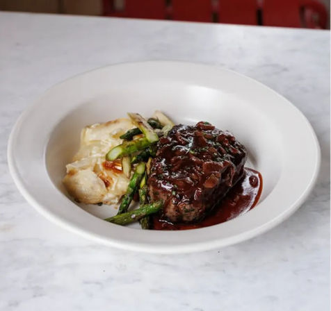 Seared steak in glossy red wine sauce with roasted asparagus and creamy potato gratin, plated in a white bowl on a marble table — restaurant-style entree.