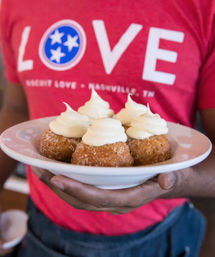 White plate of five sugar-coated doughnut holes topped with swirls of vanilla cream, held in hands against a red "LOVE Nashville, TN" shirt